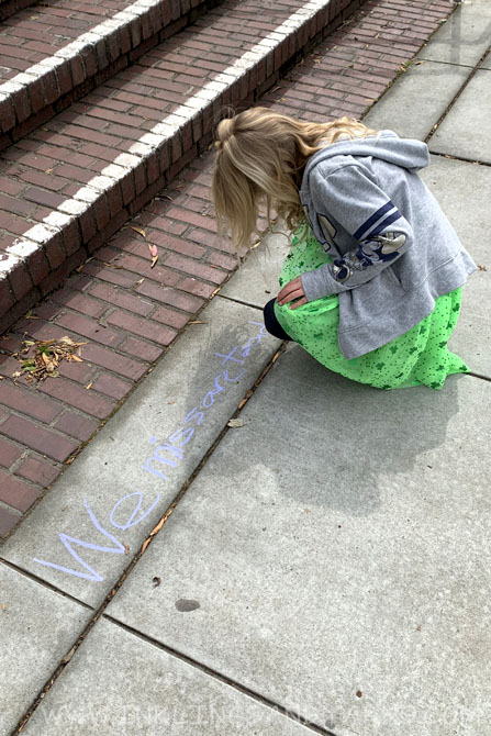 A little girl in a green tutu writing on the sidewalk outside of school.