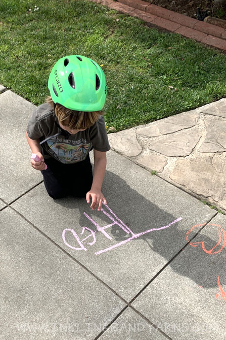 A little boy in a bike helmet writing his name in chalk on the sidewalk.